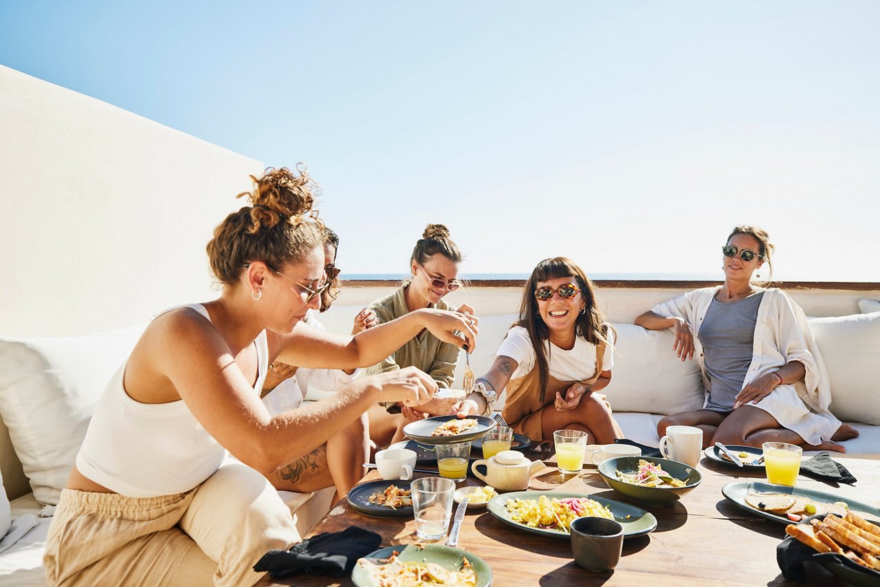 Medium wide shot of smiling female friends sharing breakfast on deck of luxury suite at tropical resort