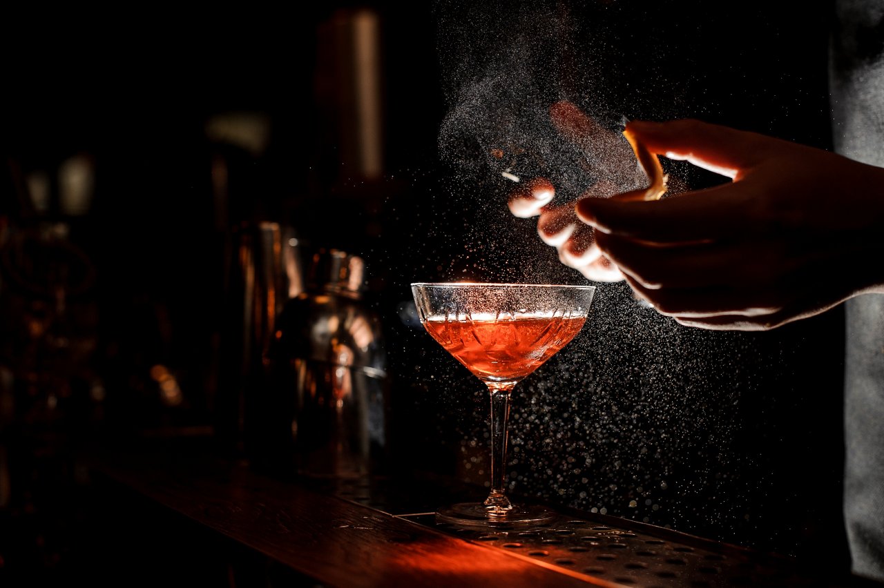 Bartender in black apron and blue shirt sprays an orange peel in cocktail glass with ice at a bar counter; Shutterstock ID 776607895; purchase_order: -; job: -; client: -; other: -